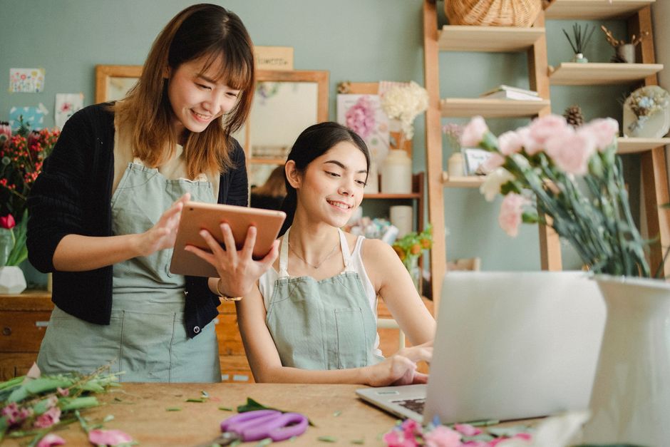 A florist with shoulder-length brown hair, wearing a light green apron over a white shirt and a black cardigan, is smiling as she displays a tablet to a seated female customer. The customer, with long