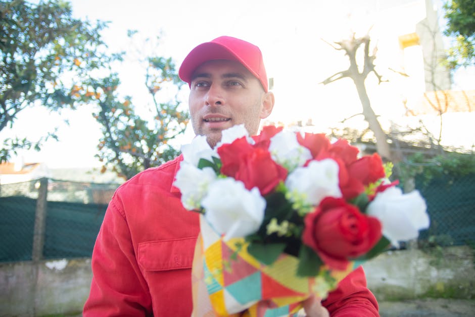 A delivery person, dressed in a red cap and matching red jacket, holding a floral bouquet wrapped in colorful paper. The bouquet features a mix of vibrant red and white roses with lush green foliage, 