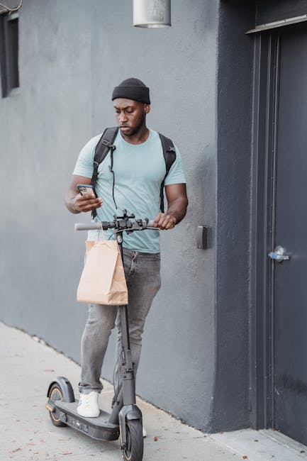 A young man with dark skin, wearing a dark beanie, light blue t-shirt, and grey jeans, stands next to a grey building wall on a grey sidewalk. He is holding a smartphone in his right hand and has a br