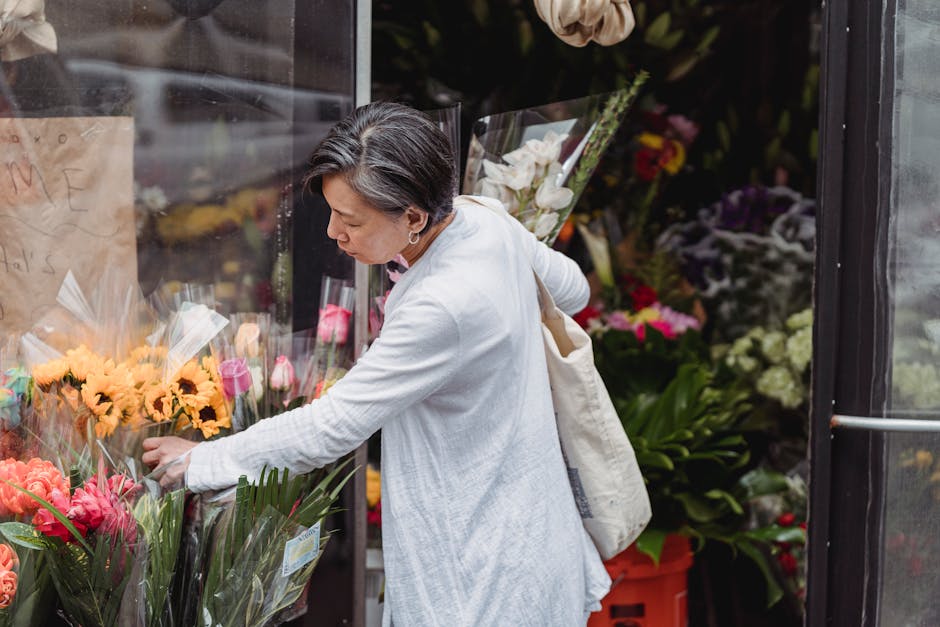 A woman with short, gray hair, wearing a white long-sleeved shirt, is selecting fresh flower bouquets from a display outside a flower shop. The arrangement features bright yellow sunflowers, pink tuli